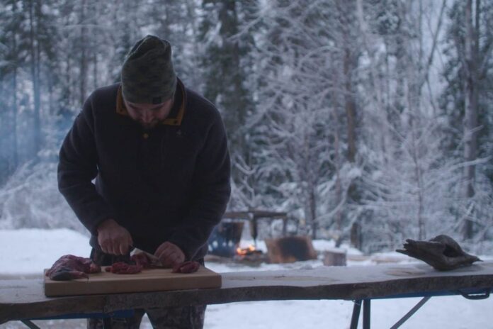 Admid a snowy background of frozen trees, a man in a hat and black jacket cut meat at an outside table.