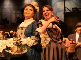 Two women of the ensemble, one in a blue 1700s dress the other in a brown and black 1700s dress.