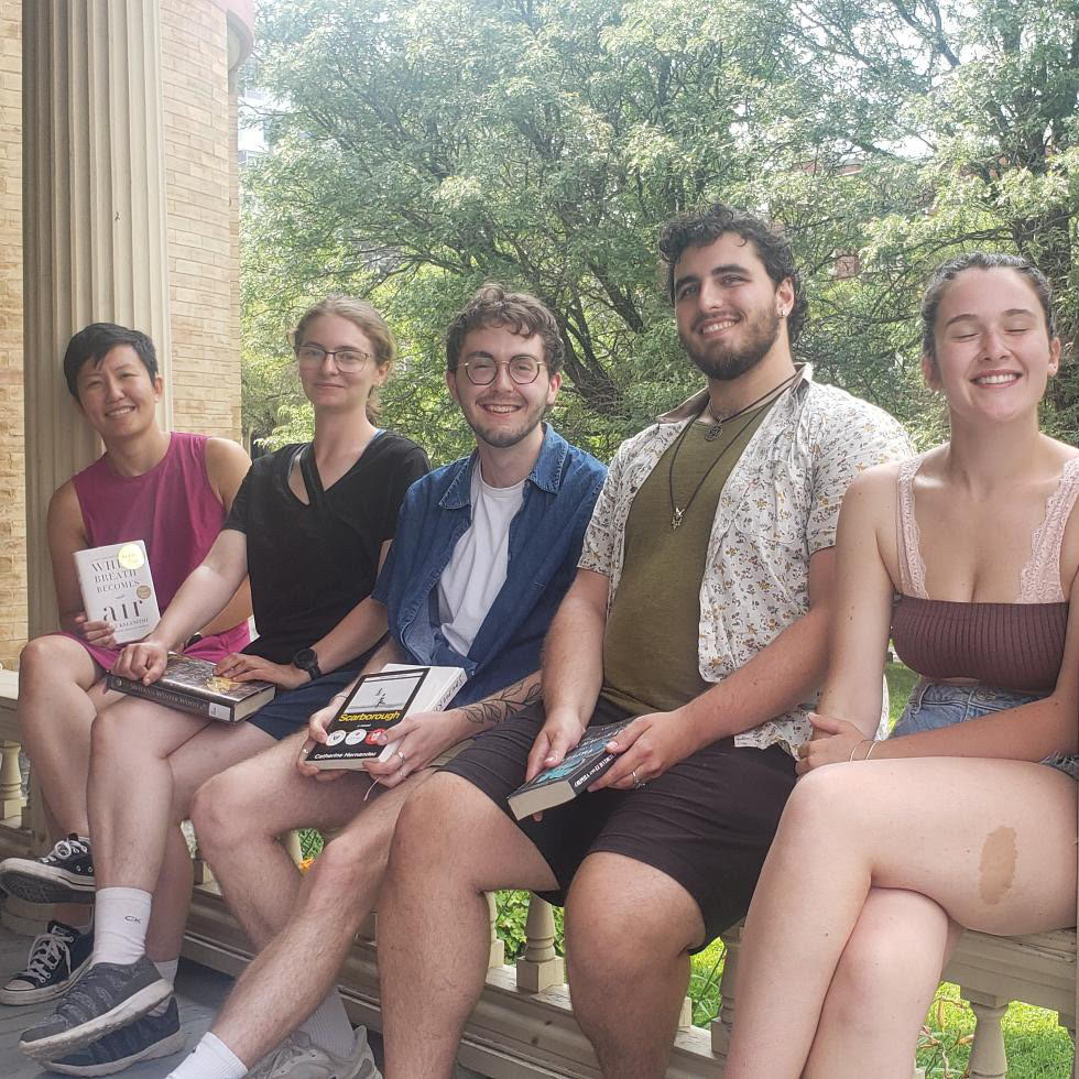 Sitting down on a balcony ledge, five people smile big while holding their book club books. 