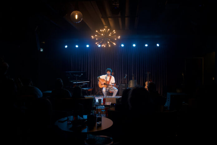 Amid a dark room, a man in a white shirt plays the guitar on stage to a crowd.