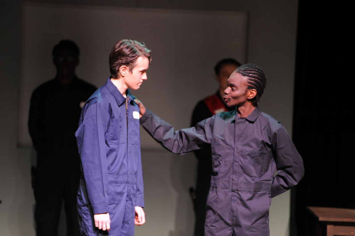 Two men in blue jumpsuits stand on stage. The one on the right has his hand on the other man's shoulder. 