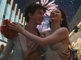 A man and a woman wearing white in an embrace with fireworks and lights in the background.