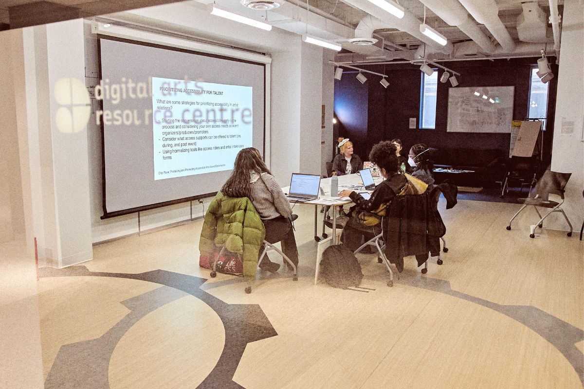 In a board meeting room, six people sit around a table with masks on a meet. They are all wearing masks and there is a projector on with text. 