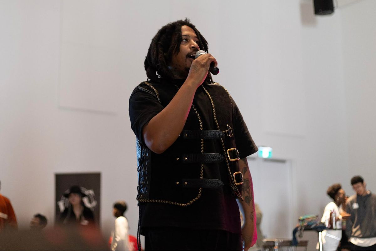 In a gymnasium, a black man wearing a black shirt holds a mic and speaks to the crowd. 