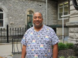 A Black man wearing a white and blue tshirt stands outside in front of a grey cobblestone building.