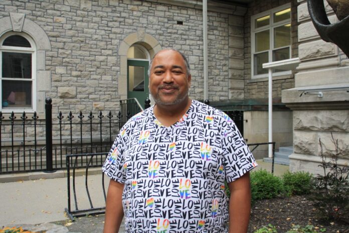 A Black man wearing a white and blue tshirt stands outside in front of a grey cobblestone building.