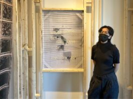 A women wearing all black with a black mask, stands to the right of an all white textile art piece.