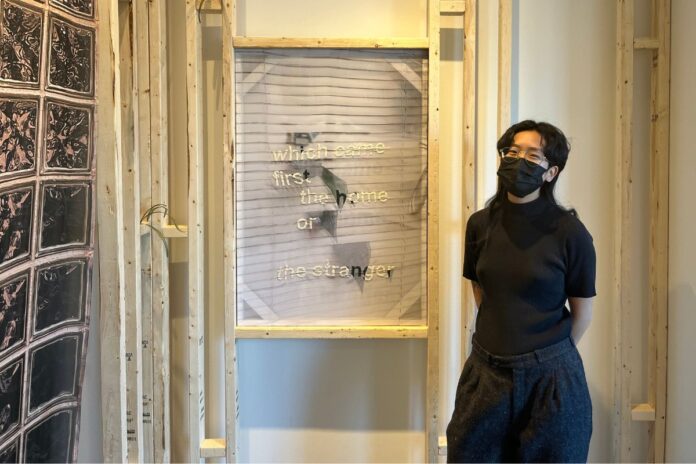 A women wearing all black with a black mask, stands to the right of an all white textile art piece.