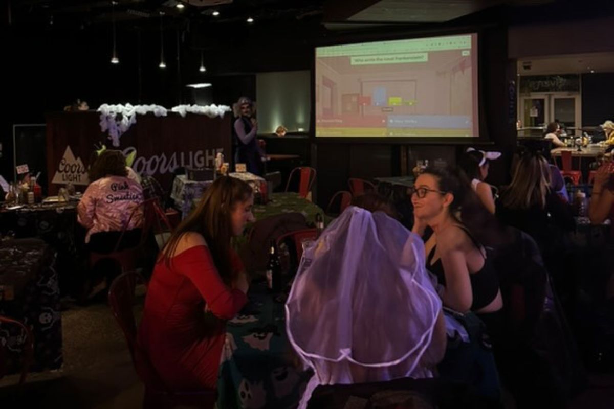 In a dark room, there is a giant white projector playing Kahoot. Sitting around small tables people are dressed up in Halloween costumes. 