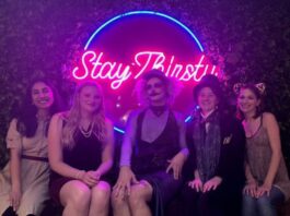 Behind a neon pink sign that says "Stay Thirsty" five people sit and smile at the camera. They are dressed in various Halloween costumes