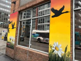 Wrapped around a brick building, a mural of a sunset depicting a geese flying over head is painted. There are also flowers (like daisy's) on the bottom of the mural