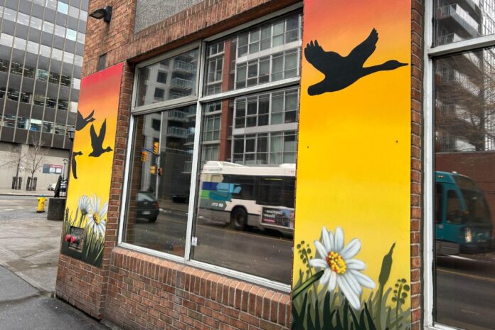 Wrapped around a brick building, a mural of a sunset depicting a geese flying over head is painted. There are also flowers (like daisy's) on the bottom of the mural