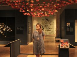 In a gallery room, a woman stands in front of her pink crocheted flowers display