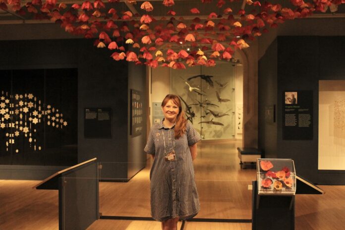 In a gallery room, a woman stands in front of her pink crocheted flowers display