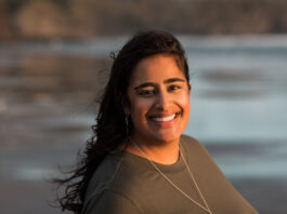 A woman with brown hair and dressed in a brown shirt smiles at the camera against a beach background.