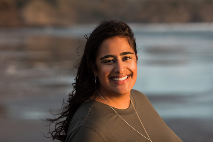 A woman with brown hair and dressed in a brown shirt smiles at the camera against a beach background.