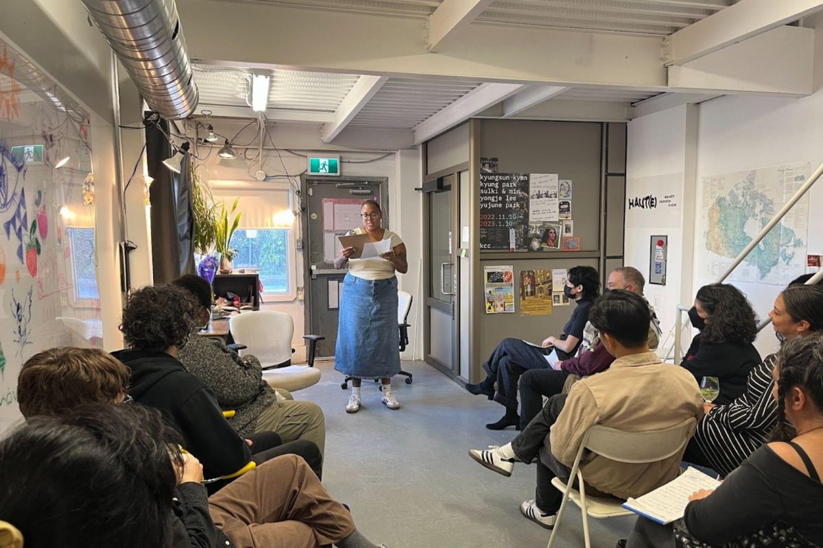 In a gallery room, a woman stands up and reads her poetry in front of a room full of around 15 people that are seated. 