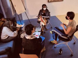 In a room, four masked people sit around a table and discuss ideas. There are notebooks out and pencils and a whiteboard.