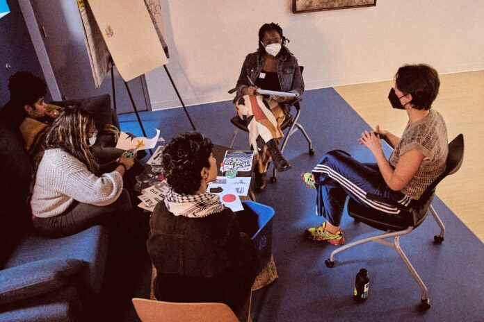 In a room, four masked people sit around a table and discuss ideas. There are notebooks out and pencils and a whiteboard.