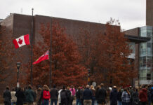 A crowd of people stand in front of the Canada and Ontario flags at half-mast.