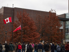 A crowd of people stand in front of the Canada and Ontario flags at half-mast.