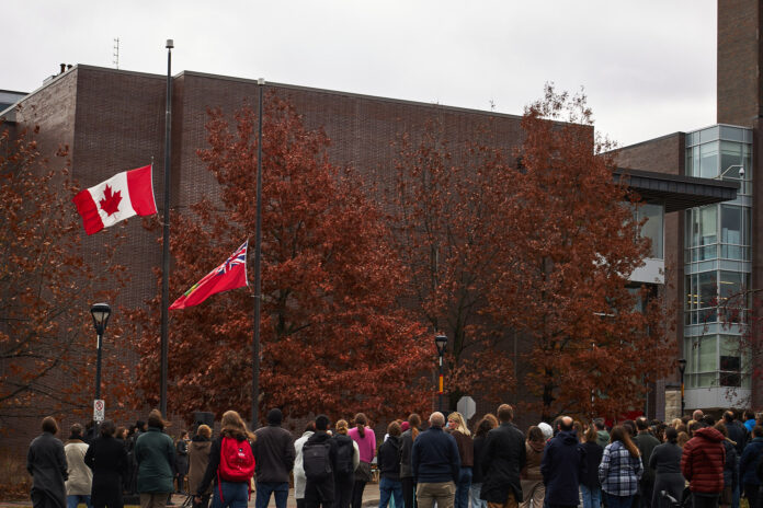 A crowd of people stand in front of the Canada and Ontario flags at half-mast.