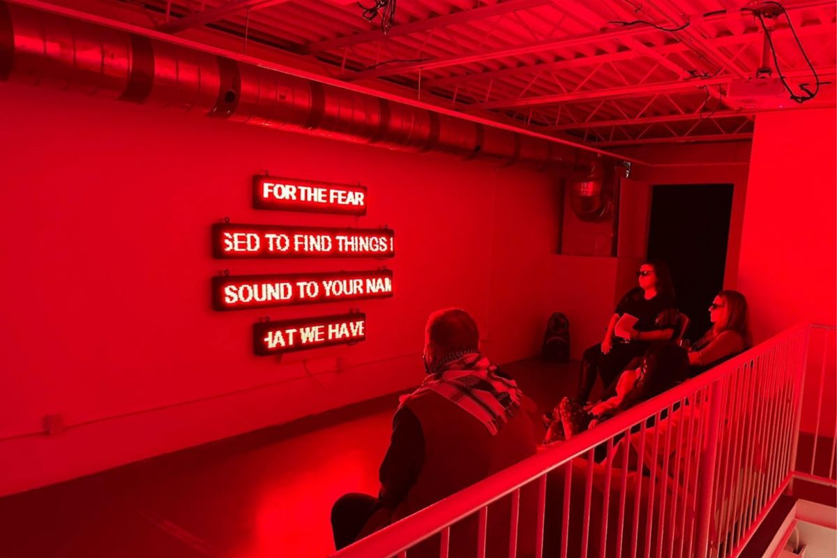 A group of attendees wearing sunglasses and sitting around the four-channel lit text. They are bathed in a red colour. 