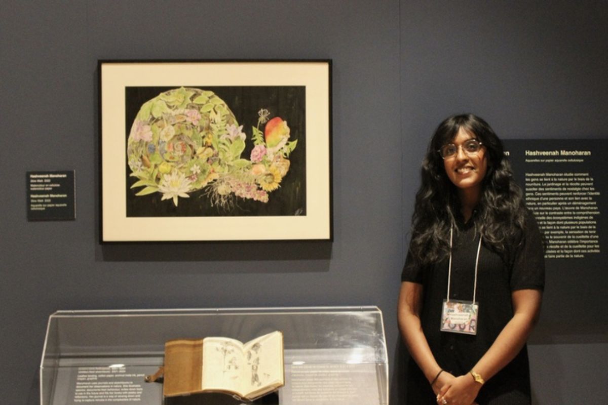 Against a grey wall, a woman stands next to her which depicts the connection between culture and the natural world