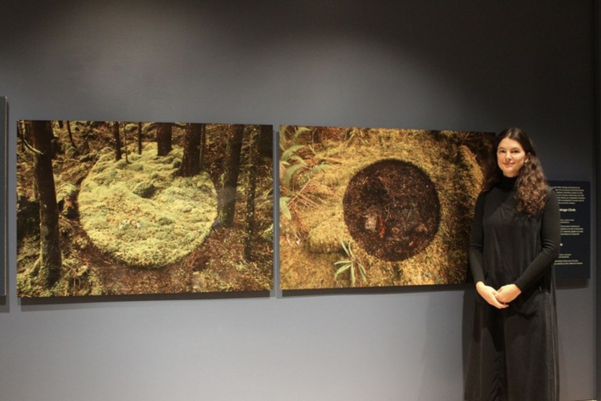 Against a grey wall, a women stands next to two photos that depict moss transplanting initiatives. 