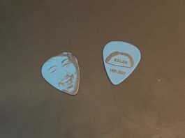 Two light blue guitar picks against a black table background. The left pick shows a sketch of Taylor’s face, while the right pick shows his nickname, “Riles,” on a taco with his date of birth and death.