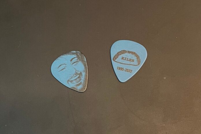 Two light blue guitar picks against a black table background. The left pick shows a sketch of Taylor’s face, while the right pick shows his nickname, “Riles,” on a taco with his date of birth and death.