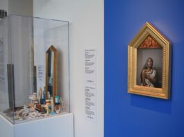 On a blue painted wall, there is a photo of a woman dressed to look like madonna. In the glass container next to it, its full of little trinkets like pill bottles, gold jewellery, and pearls.