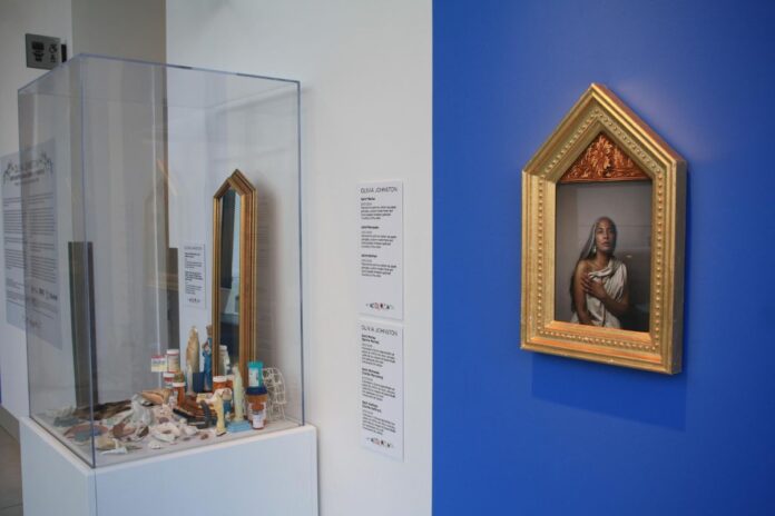 On a blue painted wall, there is a photo of a woman dressed to look like madonna. In the glass container next to it, its full of little trinkets like pill bottles, gold jewellery, and pearls.