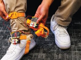 Someone wearing brown pants and white air force ones, has a tap tap device strapped to his ankle. The device has orange straps that go around the bottom of the foot and the ankle. The person shows off the battery pack and the device in one of their hands.