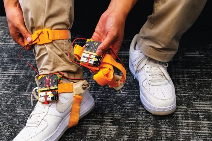 Someone wearing brown pants and white air force ones, has a tap tap device strapped to his ankle. The device has orange straps that go around the bottom of the foot and the ankle. The person shows off the battery pack and the device in one of their hands.