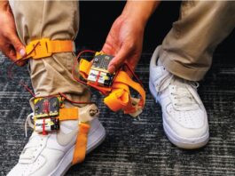 Someone wearing brown pants and white air force ones, has a tap tap device strapped to his ankle. The device has orange straps that go around the bottom of the foot and the ankle. The person shows off the battery pack and the device in one of their hands.