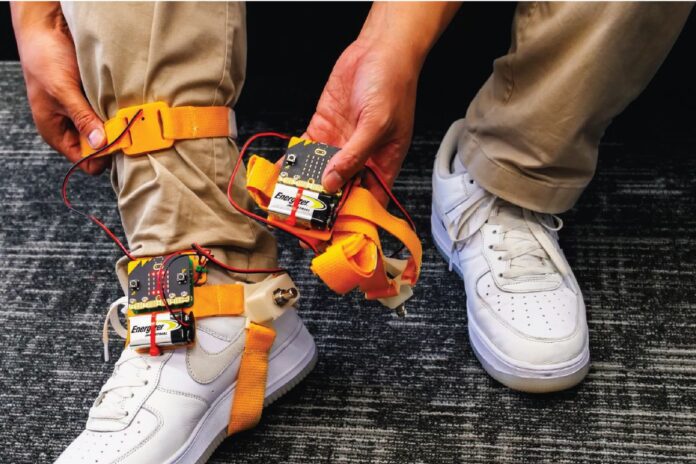 Someone wearing brown pants and white air force ones, has a tap tap device strapped to his ankle. The device has orange straps that go around the bottom of the foot and the ankle. The person shows off the battery pack and the device in one of their hands.