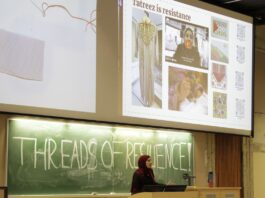 Behind a chalk board with the words "Threads of Resilience" a woman stands. On a projector above her head shows tatreez as resilience and an example of what the embroidery looks like.