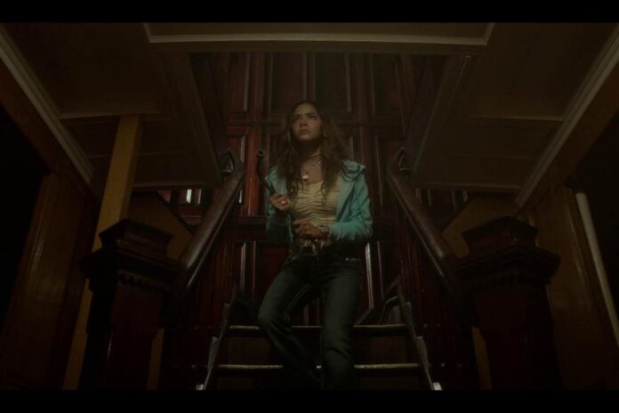 Walking down the stairs in a dark and gloomy looking basement, a girl in jeans and a white tshirt holds a knife and looks scared.