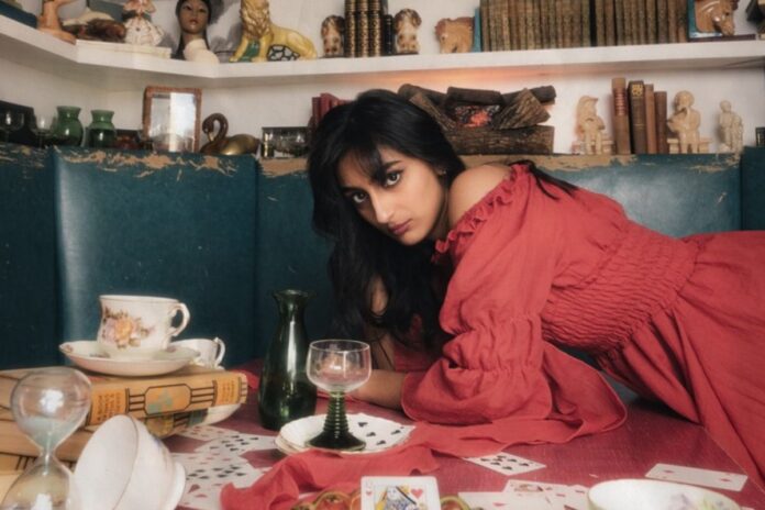 Laying on a table with teacups and cards, a person in a red dress stares directly at the camera.