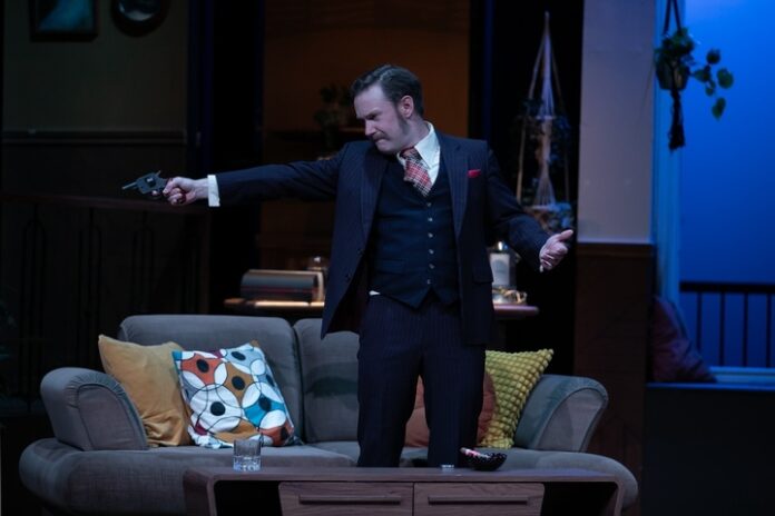 On stage, a man in a black suit points a gun to someone off stage.