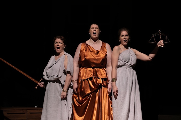 On stage, three woman dressed in white and orange togas sing with swords. 