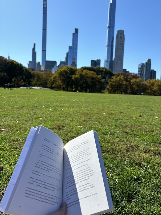 Someone holds out a book with the backdrop of the NYC skyscrapers. This person is laying in Central Park.