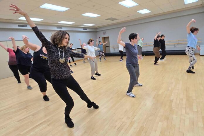 A group of dancers, all women and men in their mid to late 30s, dance with their arms out stretch while sliding to the right.