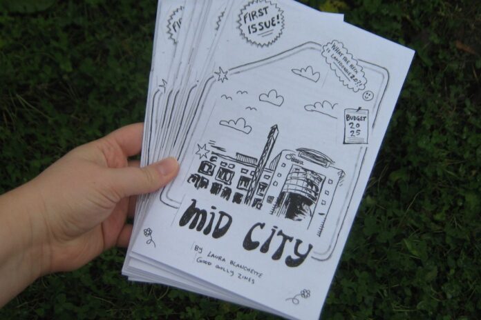 Amid a green background of grass, a hand holds out a white zine that has a drawing of buildings and has the title 