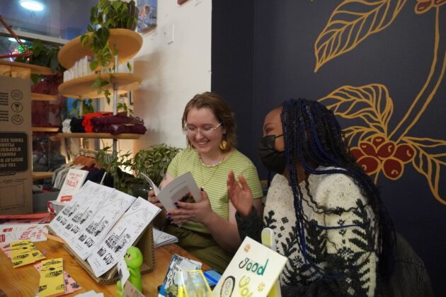 Two women laughing while holding a white small zine. They are seated and in front of them are lined up versions of the zine.