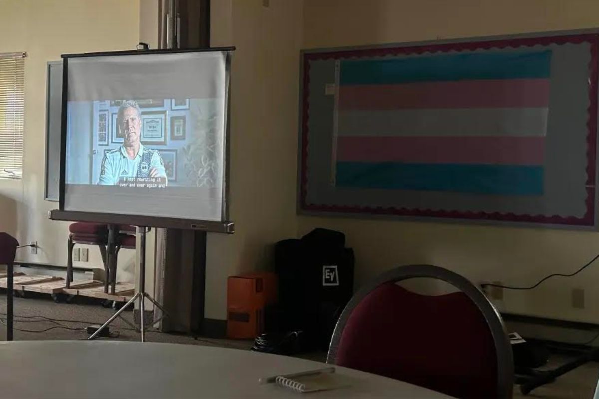 On a projector in a classroom, the movie ‘Will & Harper' is playing. There is a transgender flag hanging next to it. 