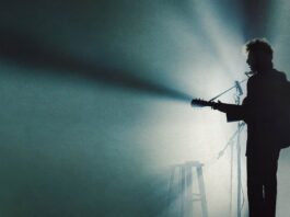 A black silhouette of a artist playing a guitar amid blue stage lighting.