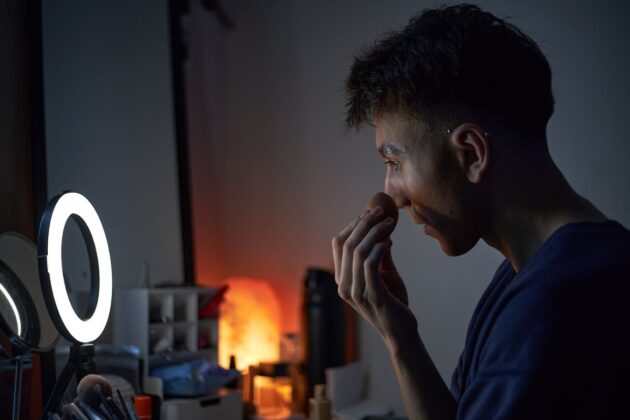 A drag queen sits in front of a vanity mirror and puts makeup on her face.