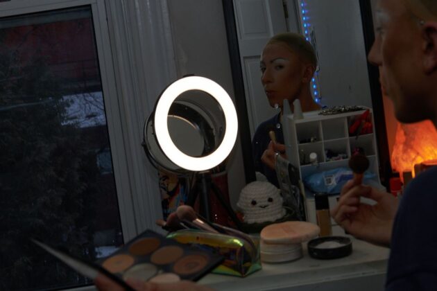 A drag queen sits in front of a vanity mirror and puts makeup on her face.